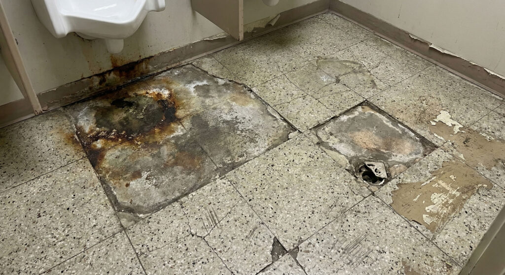 Damaged public restroom floor with corrosion and surface breakdown caused by moisture intrusion, chemical exposure, and biological contamination