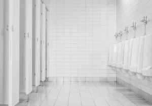 Public restroom interior with urinals, tiled walls, and stall partitions highlighting surfaces commonly exposed to moisture, bacteria, and constant foot traffic.